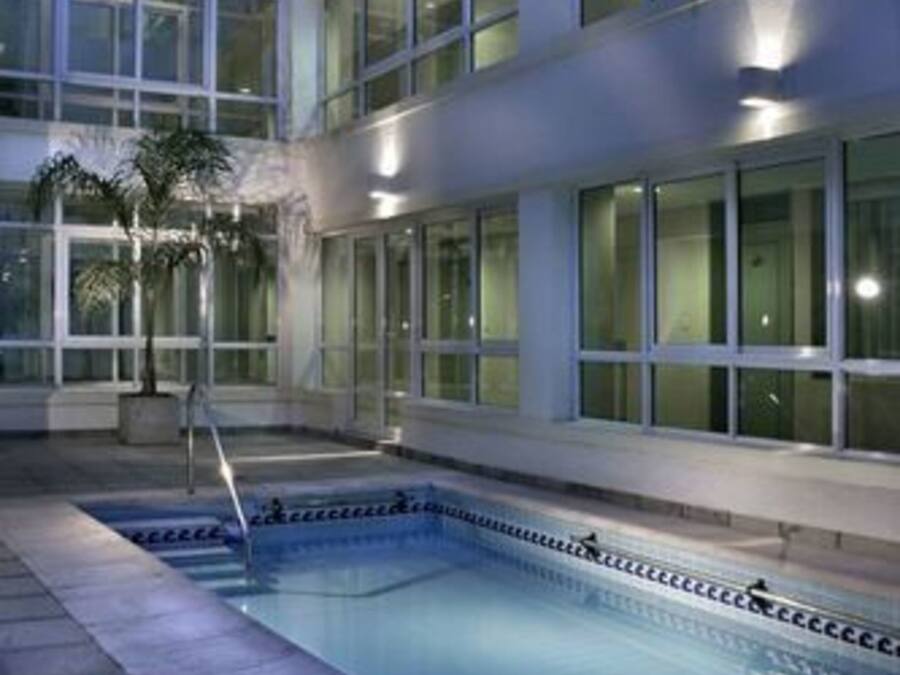 Una piscina amb un solàrium amb rajoles, envoltada per un edifici amb grans finestres i una palmera.