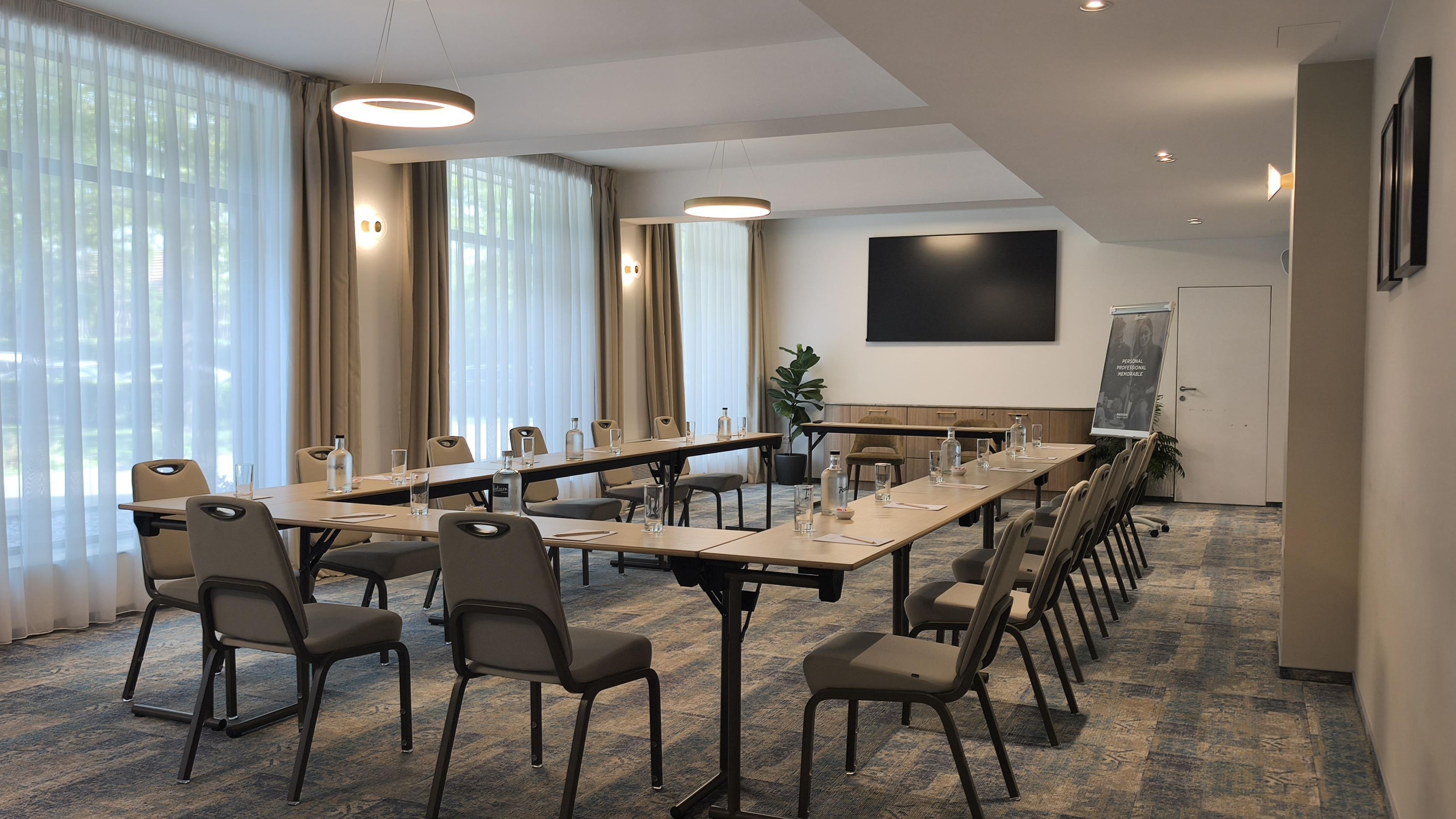 Ein Konferenzraum mit langem Tisch, Stühlen, einer Projektionsleinwand und einem großen Fenster mit Vorhängen.