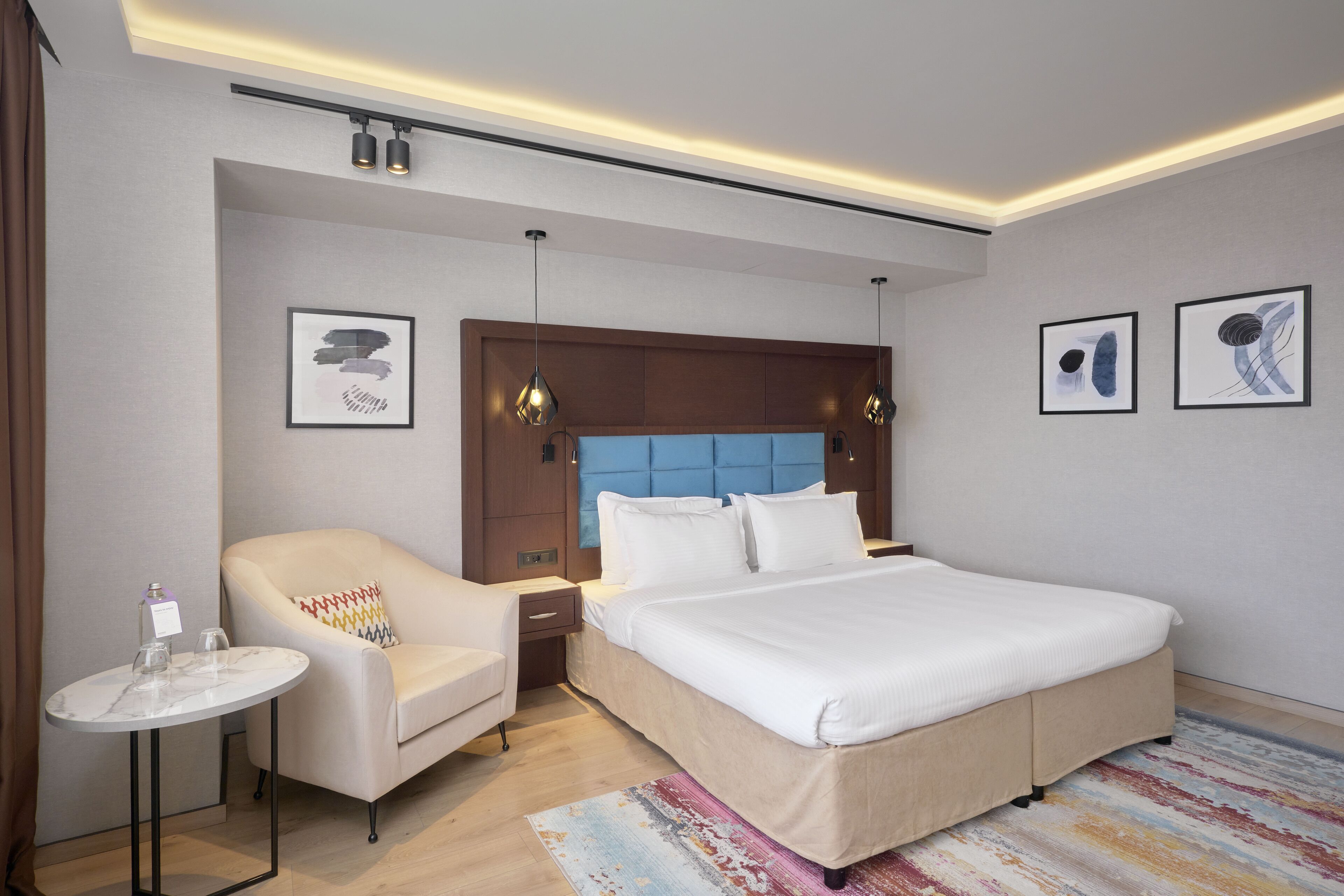 A modern hotel room with a large bed, a small round table, a sofa, and two framed artworks on the walls.