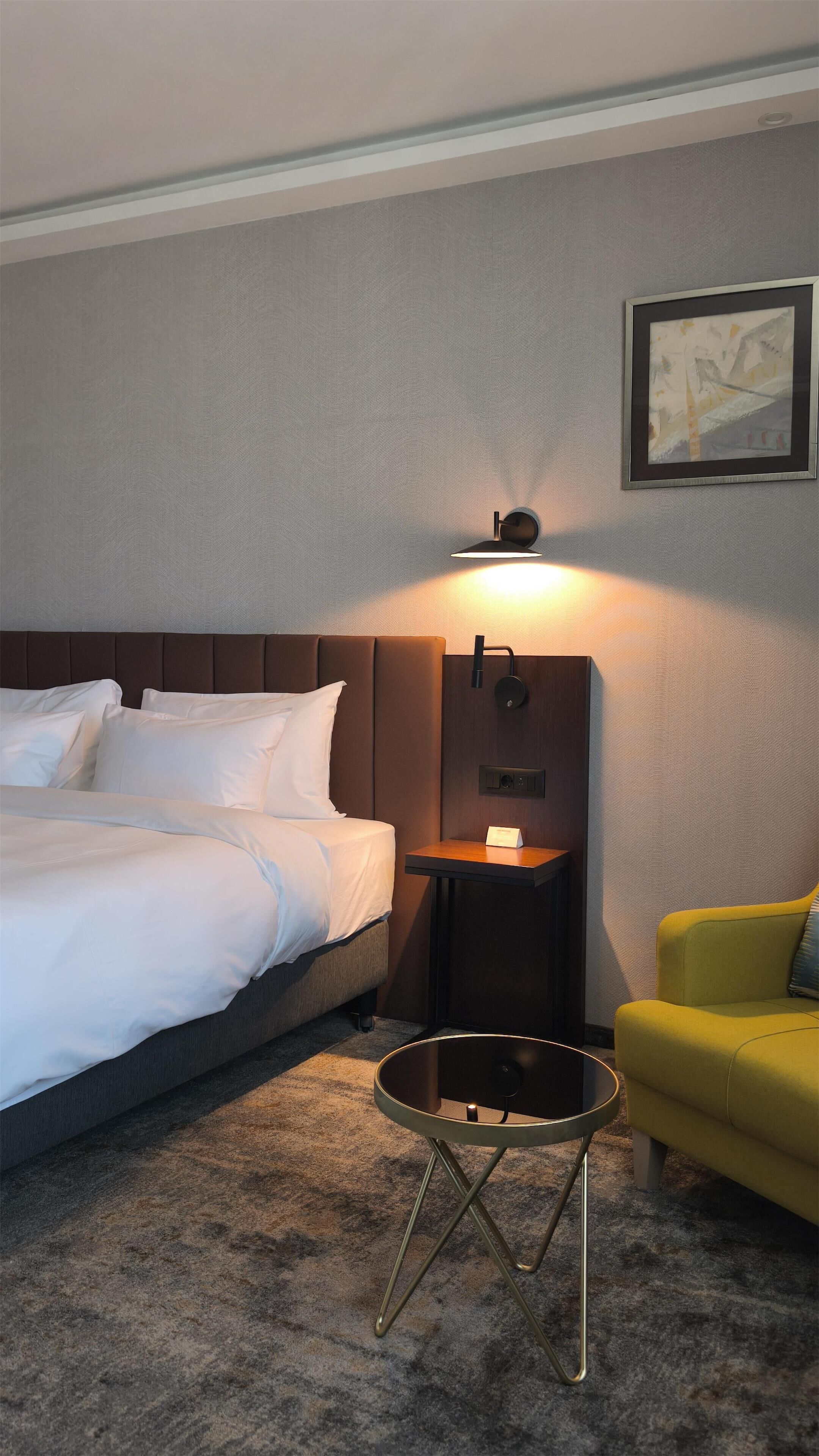 A hotel room with a bed, bedside table, lamp, and a yellow armchair.