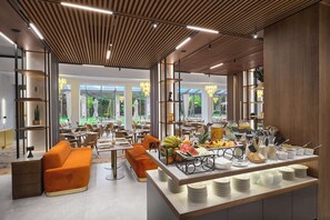A modern dining area with a buffet, orange seating, and large windows offering a view of greenery. - Radisson Hotel Plovdiv (Plovdiv)