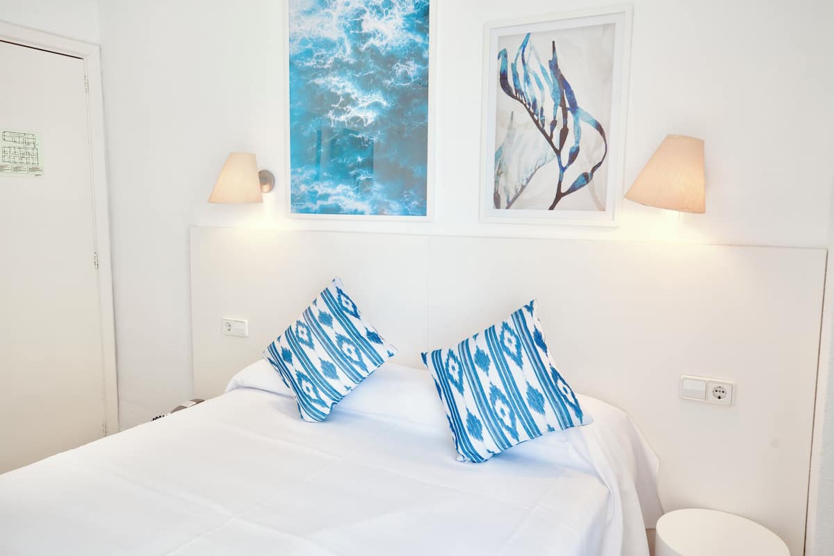 A neatly made bed with blue patterned pillows, a framed abstract painting, and two wall-mounted lamps.