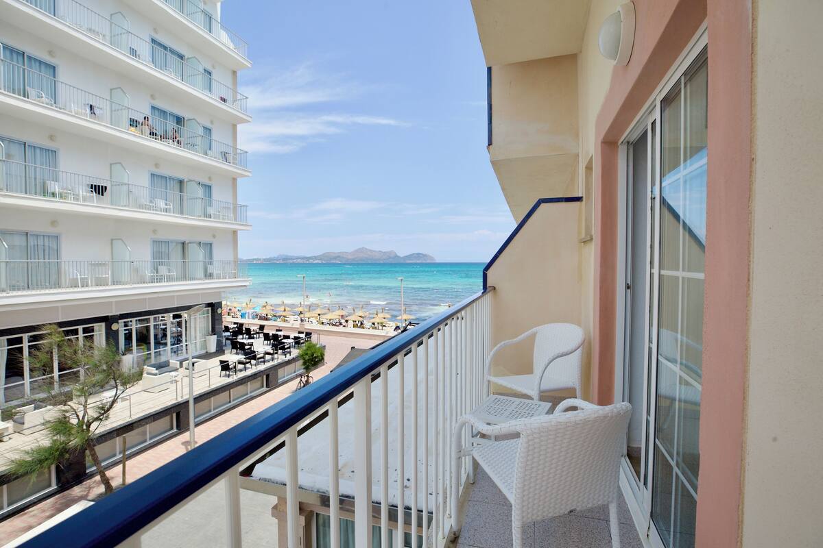A balcony with a view of the beach and ocean, white wicker furniture, and a building with multiple floors and balconies.
