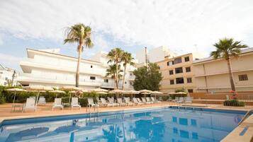Una piscina con sillones de descanso y sombrillas, rodeada de edificios y palmeras.