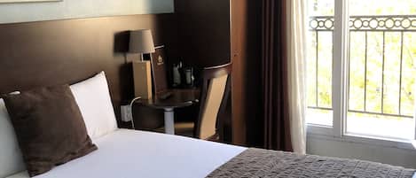 A hotel room with a large bed, a desk with a lamp, a television, and a balcony with curtains.