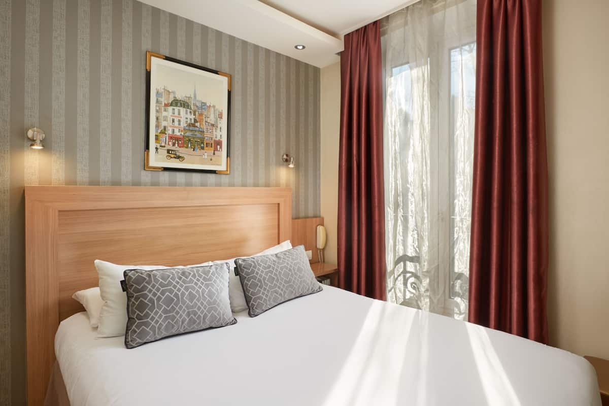 A hotel room with a large bed, a headboard with a cityscape painting, two bedside lamps, and a window with red curtains.