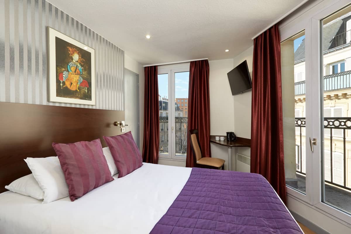 A hotel room with a bed, a desk, a chair, a TV, and a balcony with a view of buildings.
