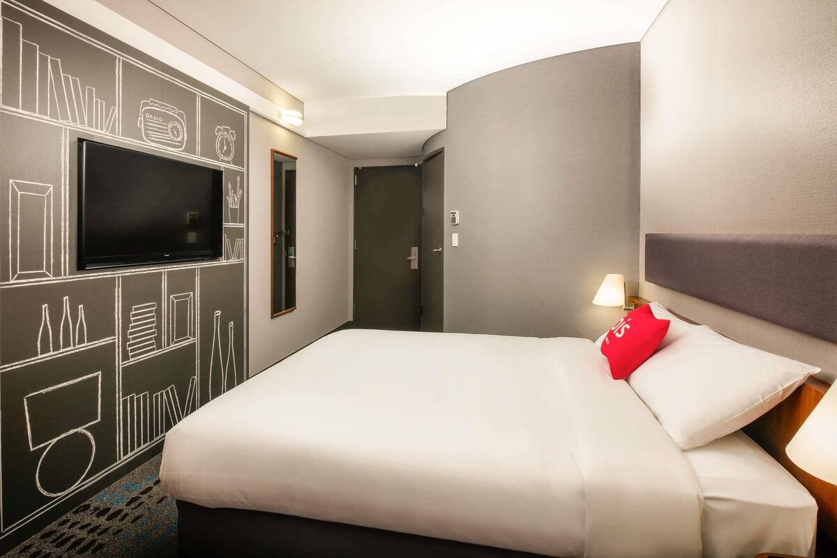 Une chambre d’hôtel comprenant un lit, un téléviseur à écran plat fixé au mur, une porte et un mur orné de dessins abstraits.