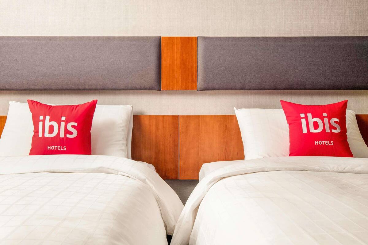 Ein Hotelzimmer mit zwei Betten, jeweils mit weißer Tagesdecke und einem roten Kopfkissen mit dem Aufdruck 'ibis HOTELS'. Das Kopfteil ist aus Holz mit einer grauen Platte.