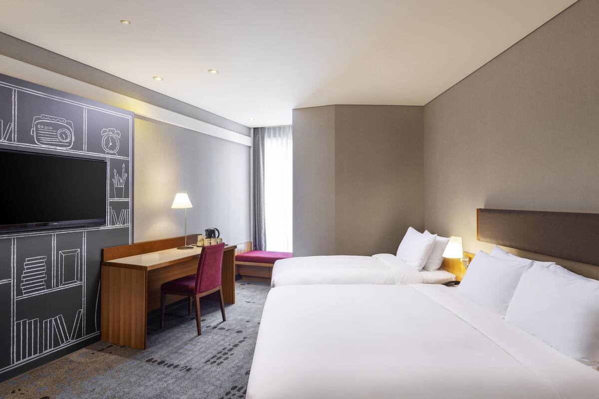 Ein Hotelzimmer mit Schreibtisch, zwei Betten, einem Fernseher und einem Fenster mit Vorhängen.