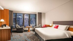 A hotel room with a large bed, a desk with a lamp, a chair, a small table, and a view of the cityscape through large windows. - ibis Ambassador Seoul Myeongdong (Seoul)