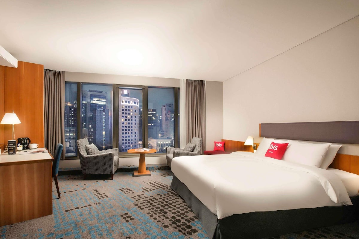 Ein Hotelzimmer mit einem großen Bett, einem Schreibtisch mit Lampe, einem Sessel, einem kleinen Tisch und Blick auf die Skyline durch große Fenster.