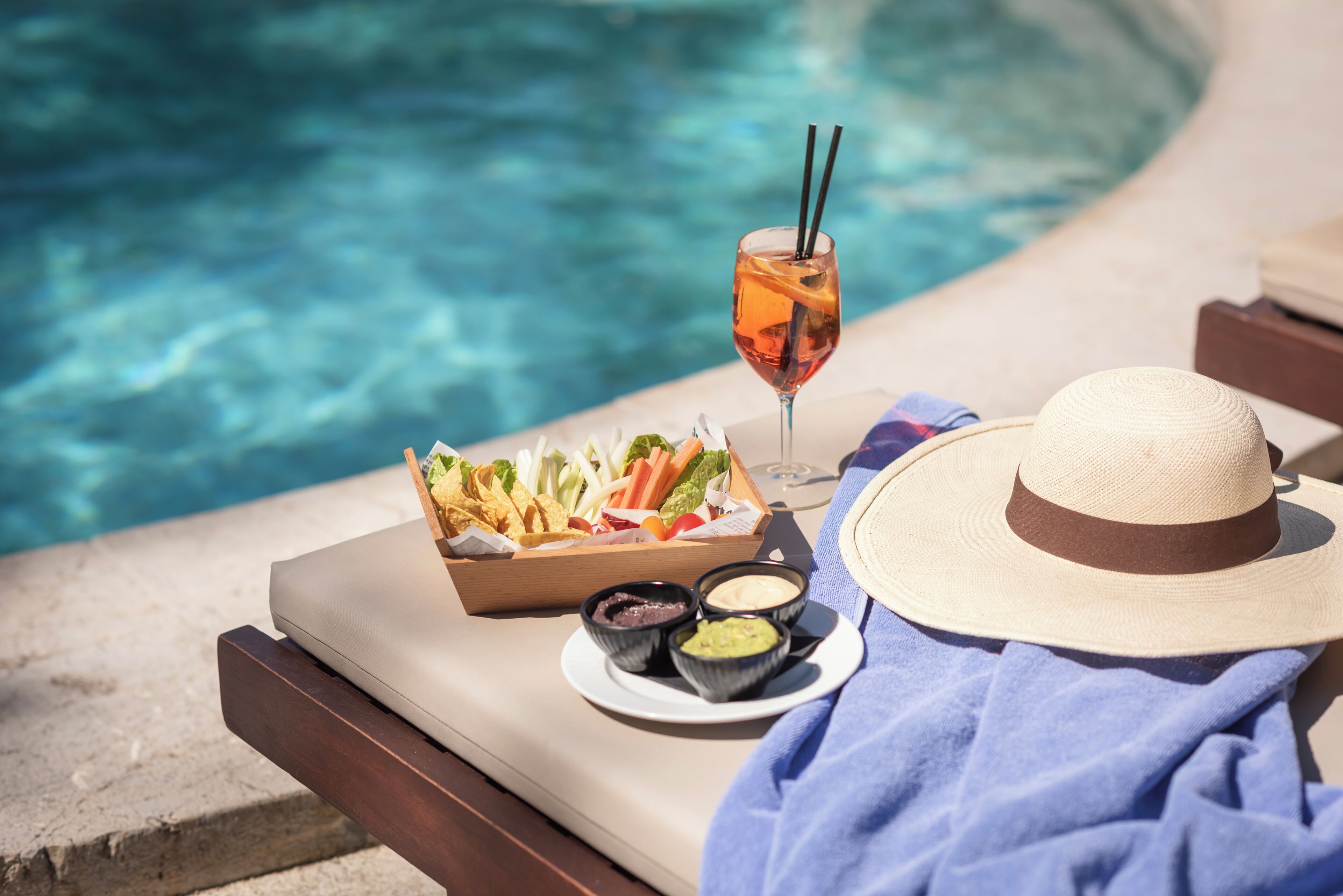 Escena junto a la piscina con una bandeja de aperitivos, una bebida y un sombrero.