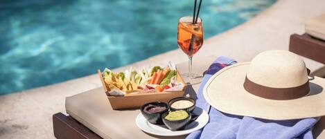 Escena junto a la piscina con una bandeja de aperitivos, una bebida y un sombrero.