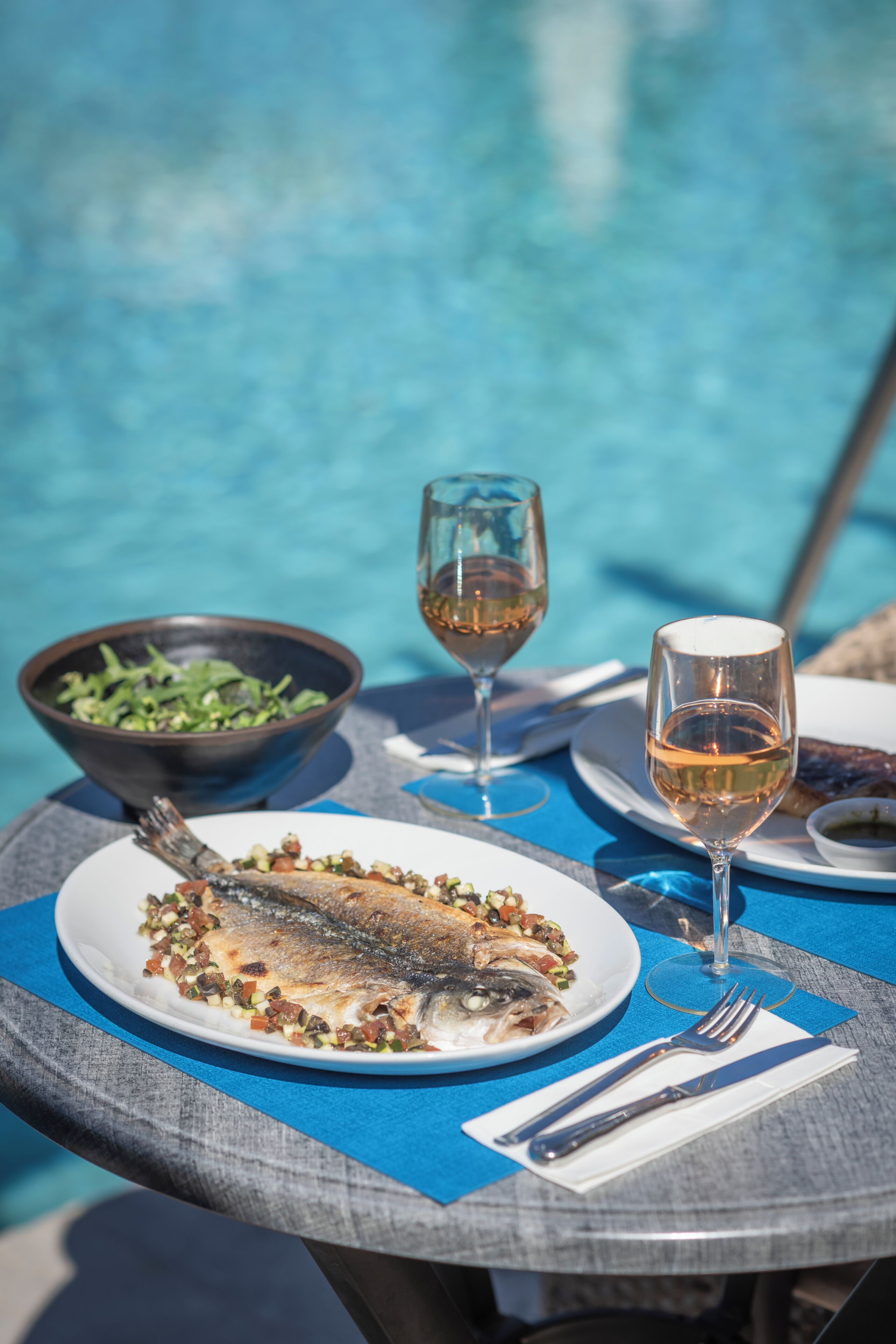 A dining table with a plate of fish, a bowl of salad, and two glasses of wine by a pool.