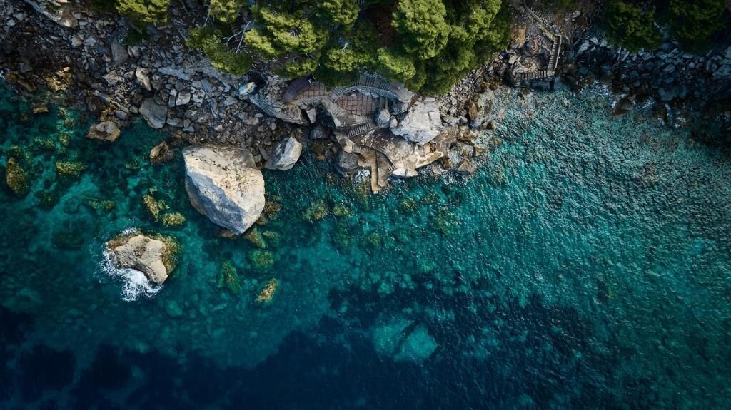 An aerial view of a coastal area with clear turquoise waters, rocky terrain, and a stone pathway leading to a small structure.