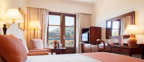 A hotel room with a bed, two chairs, a desk, a television, and a large window offering a view of a hill.