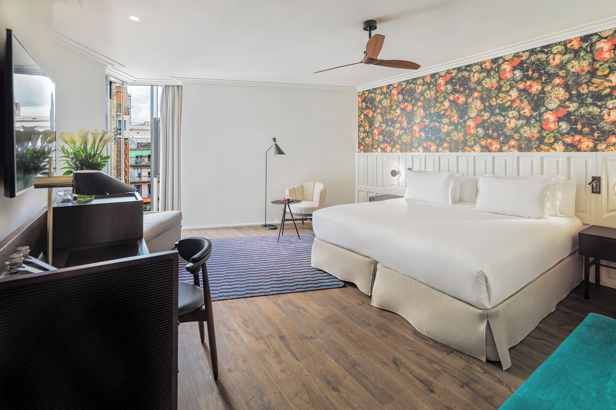 Une chambre d’hôtel avec un grand lit, un bureau, une chaise, un ventilateur de plafond et un papier peint à motifs floraux.