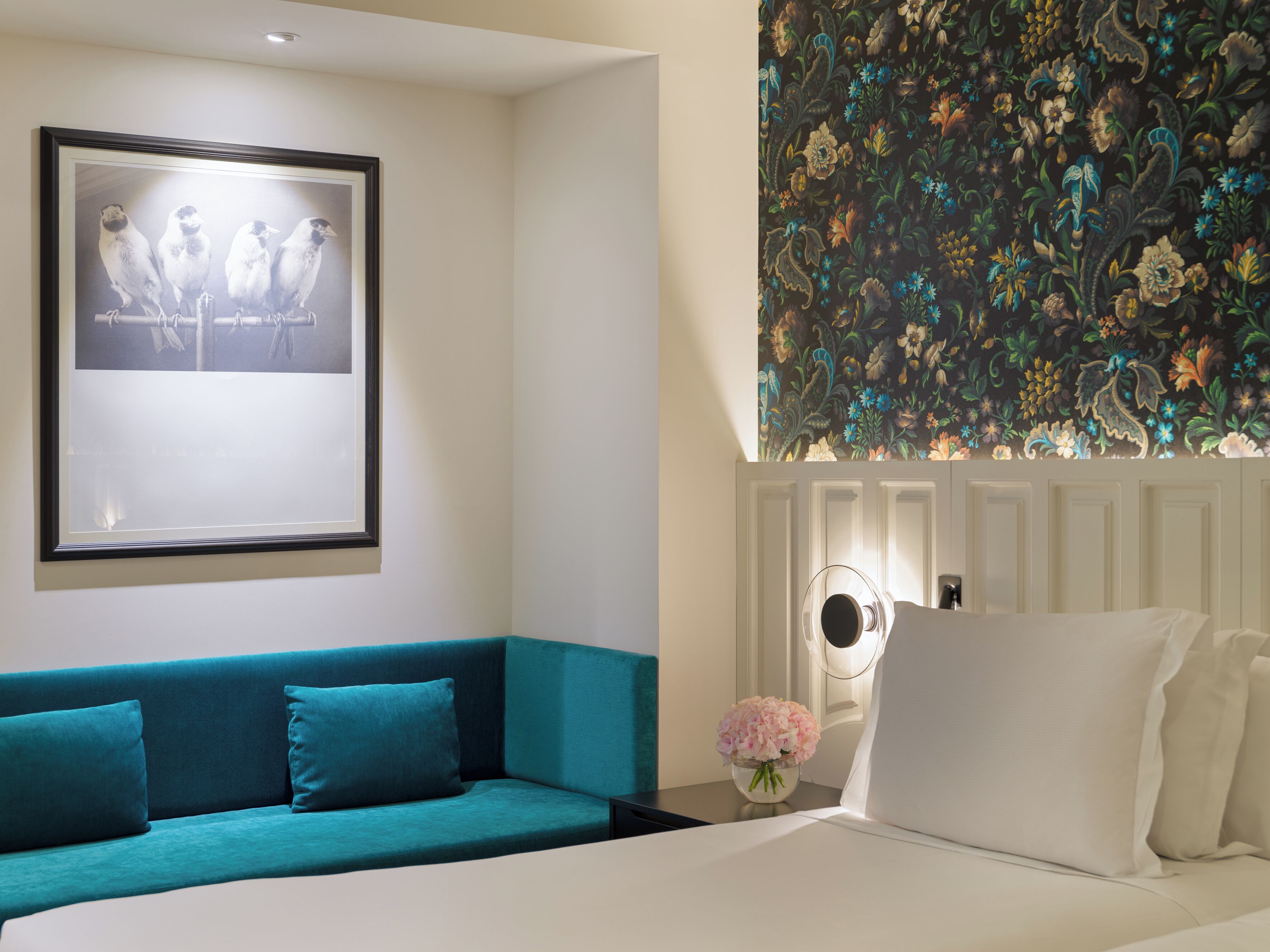 Una habitación de hotel con un sofá color turquesa, una cama con ropa blanca, un cuadro en la pared y un papel tapiz estampado con flores.