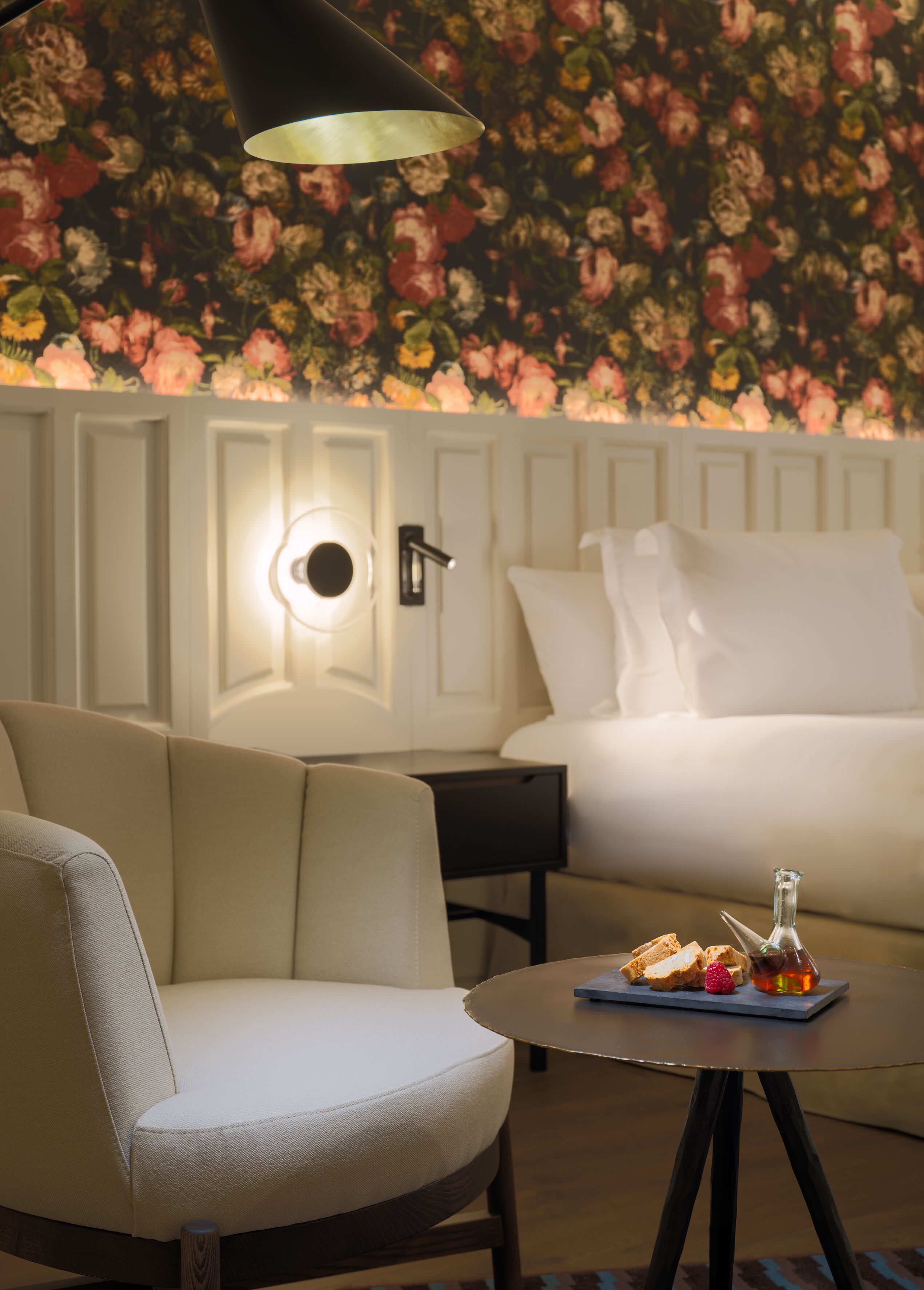 Habitación de hotel con papel tapiz floral, una cama con ropa de cama blanca, una mesita de noche con una lámpara, una silla y una pequeña mesa con una botella y un vaso.