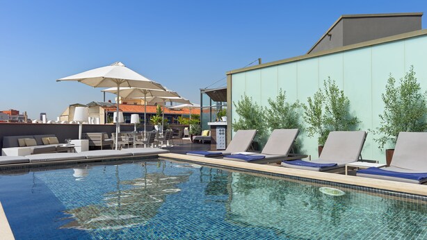 Un espace piscine sur le toit, avec des chaises longues et des parasols, un coin salon avec des canapés et une vue sur le paysage urbain.