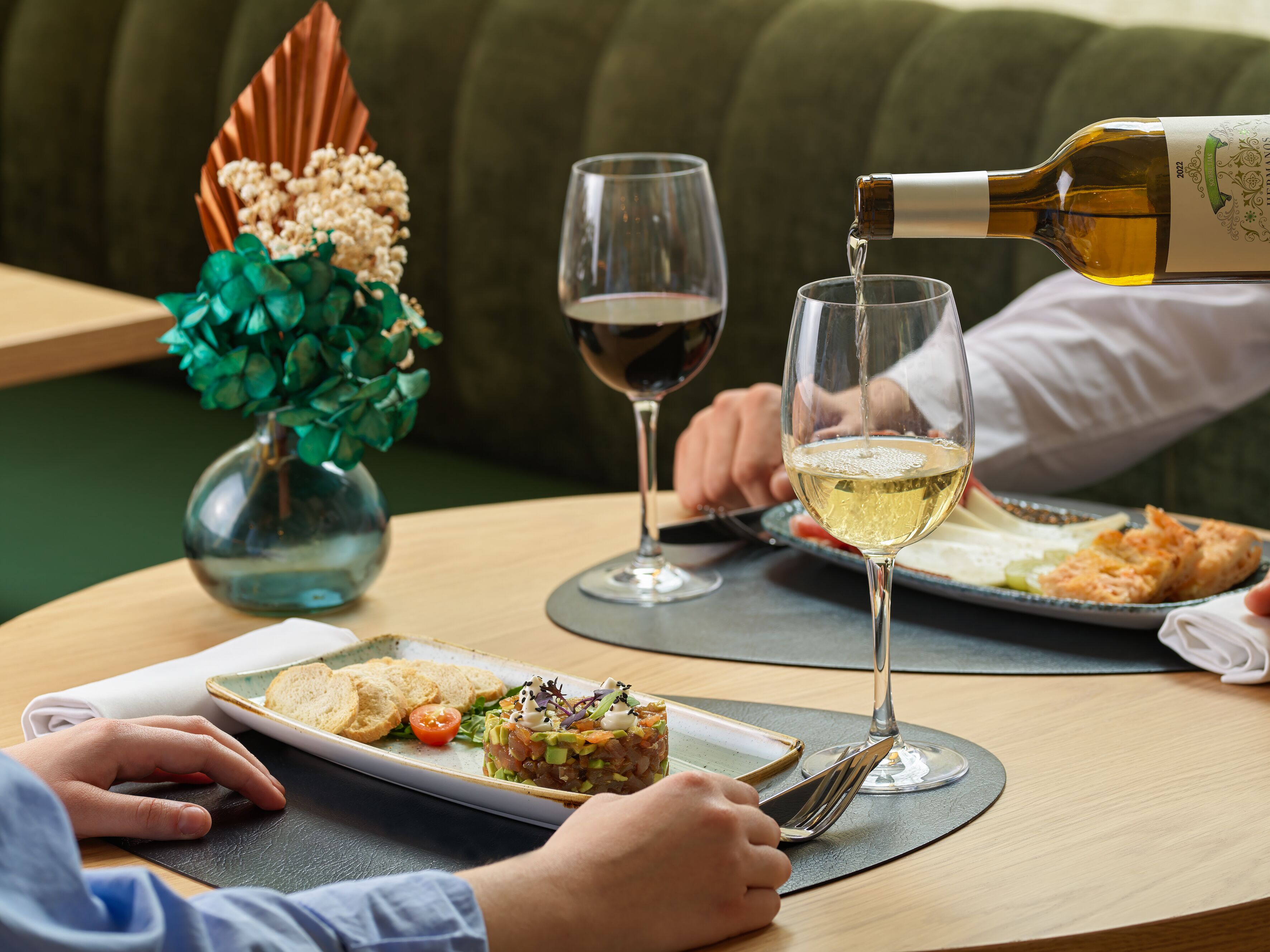 Une table à manger avec une assiette de nourriture, un verre de vin blanc et une bouteille de vin dont on verse le contenu.