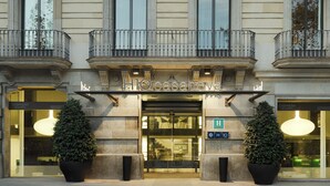 La façade d’un hôtel présente un style architectural classique, avec une entrée imposante dotée d’une porte en verre, deux plantes en pot de chaque côté et un balcon orné de balustrades.