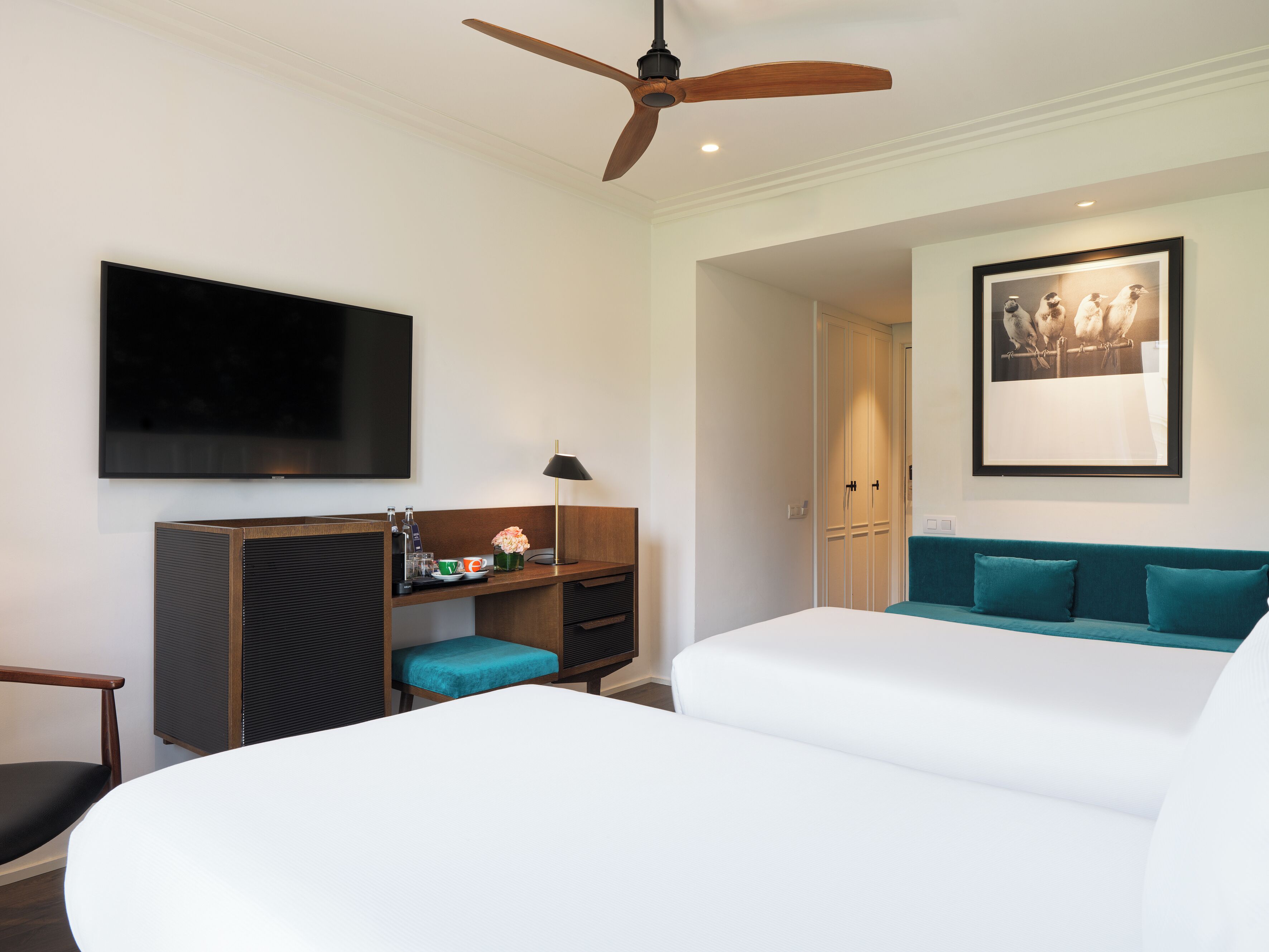 Habitación de hotel con cama, escritorio con computadora, televisor, ventilador de techo y un cuadro en la pared.