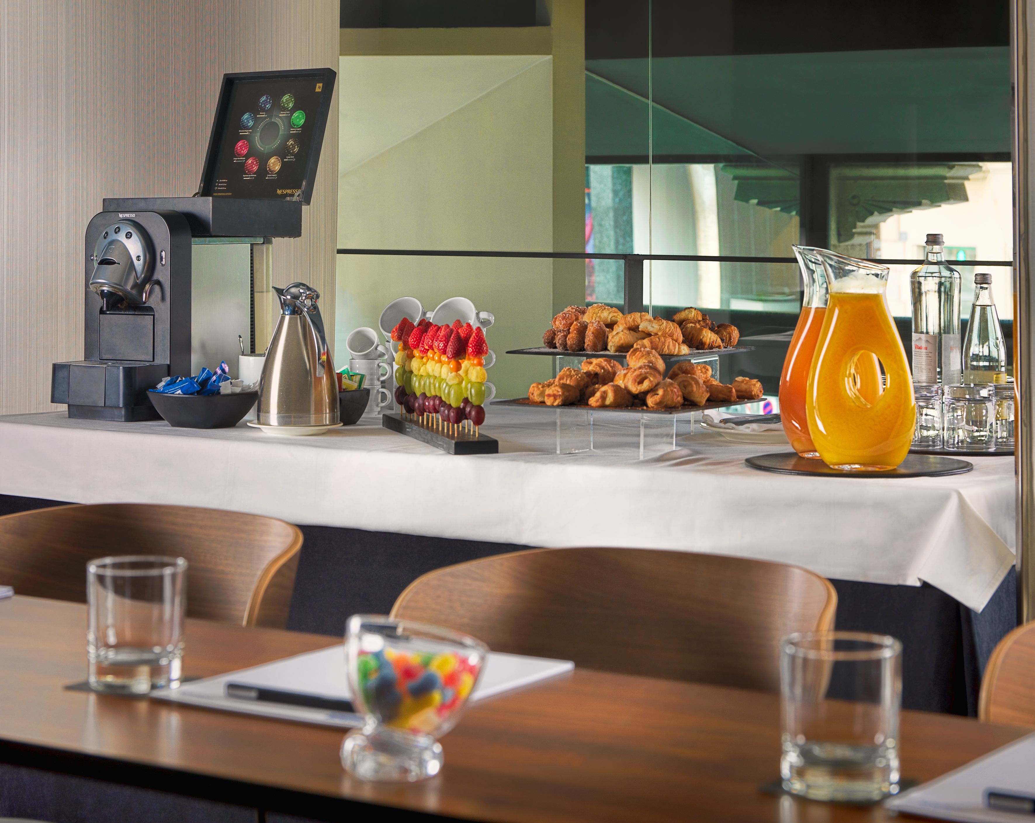 A breakfast buffet with a coffee machine, fruit skewers, pastries, and a pitcher of orange juice.