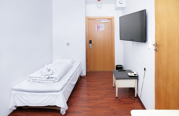 Een kleine, schone kamer met een enkel bed, een bureau, een stoel, een televisie en een deur.