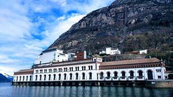 Un edificio histórico con fachada blanca y ventanas arqueadas, situado junto a una masa de agua con montañas al fondo.