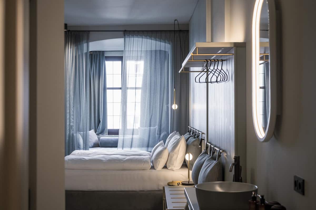 Habitació d'hotel moderna amb un llit gran, un mirall rodó, un tocador integrat i una finestra amb cortines translúcides.