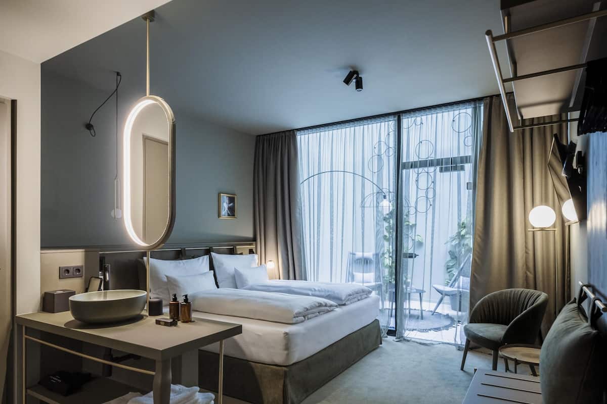 Una habitació d'hotel moderna amb un llit gran, un escriptori amb un mirall, una cadira i vistes a l'exterior a través de grans finestres.