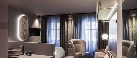 Una habitación de hotel moderna con una cama grande, un escritorio con espejo, un otoman redondo y una zona de estar con sofá y sillones.