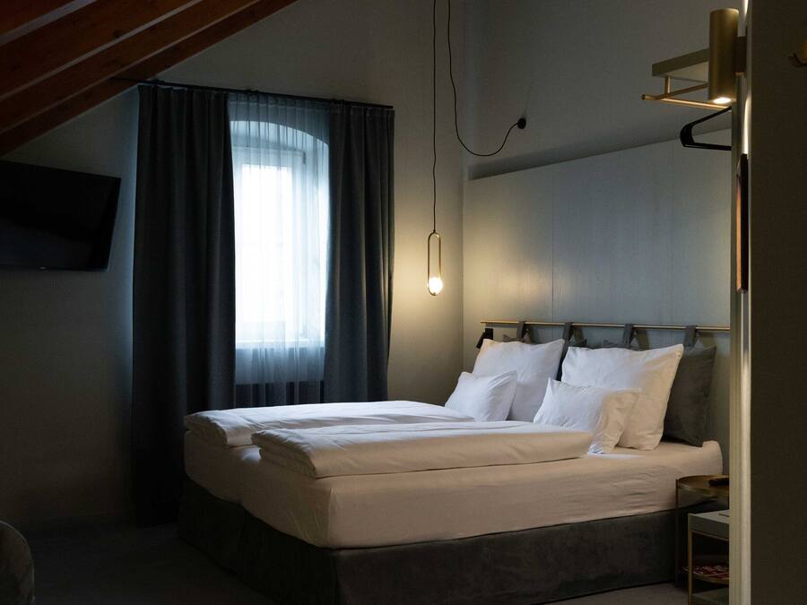 A bedroom with a bed, a bedside table, a wall-mounted TV, and a window with curtains.