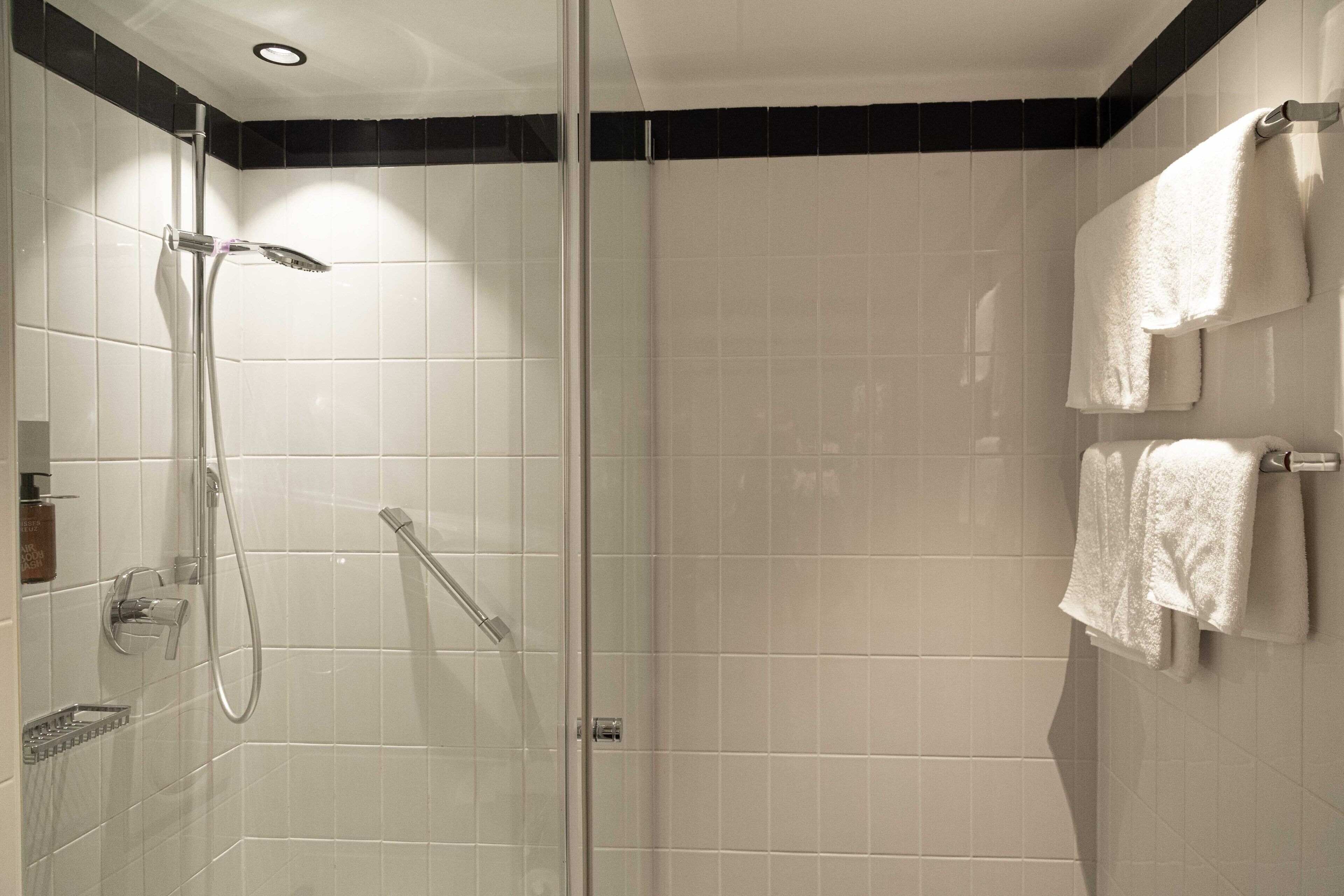 Un baño moderno con ducha envuelta en cristal, azulejos blancos y dos toallas colgadas en un perchero.