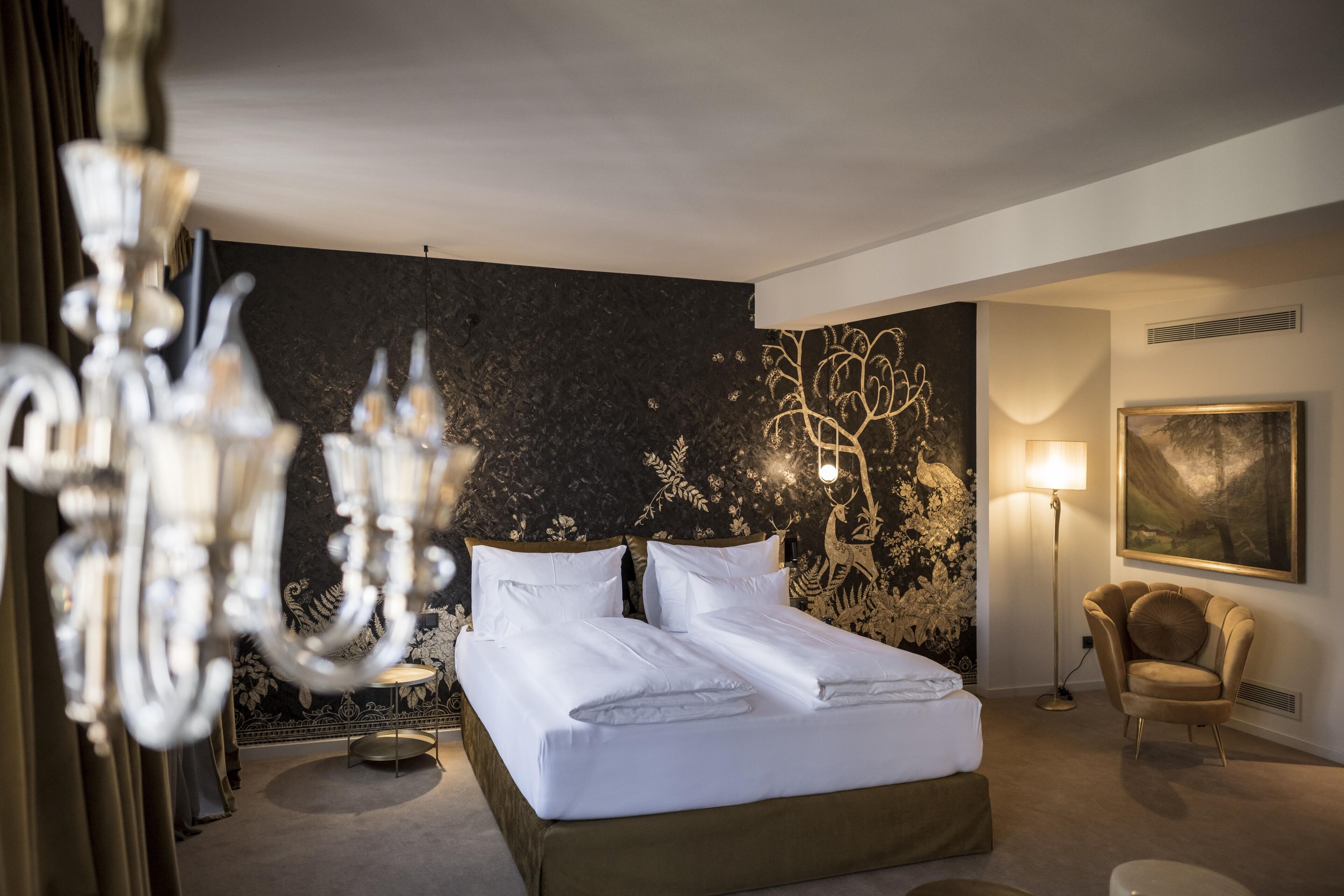 Une chambre d’hôtel avec un lit, un lustre, un mur orné de motifs floraux, une lampe et une chaise.