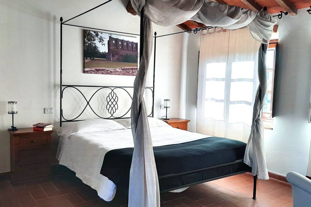 A four-poster bed with a canopy, a nightstand with a lamp, a framed picture on the wall, and a window with curtains.