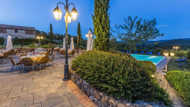 An outdoor dining area with stone tables and chairs, a pool, and a stone building in the background. - Poggio Cennina Resort (Bucine)