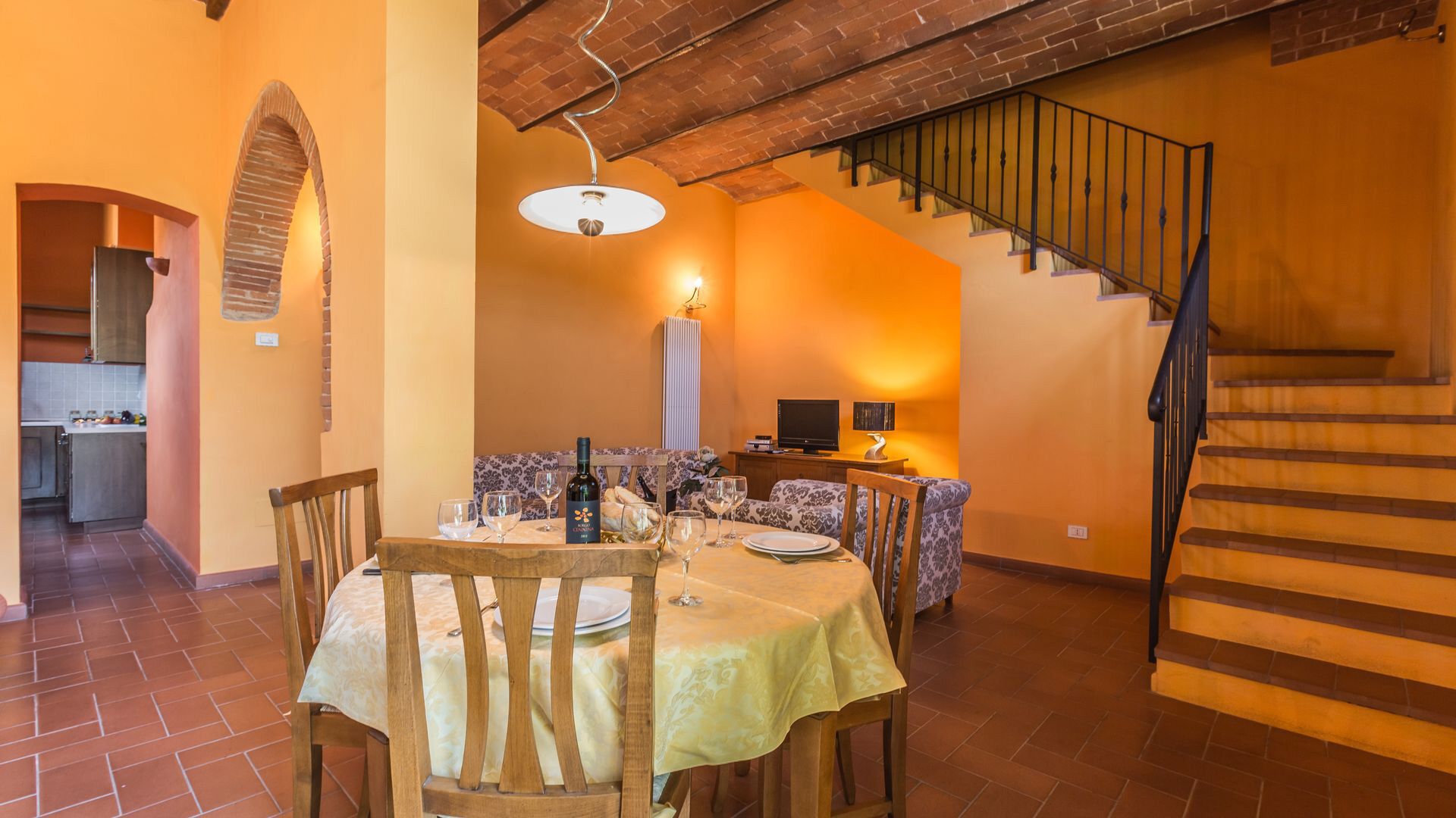 A dining area with a table set for two, a kitchen in the background, and a staircase leading to an upper level.