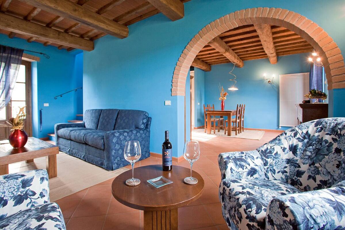 A living room with a blue sofa, a floral-patterned armchair, a wooden coffee table with two wine glasses and a bottle, and an arched brick structure.