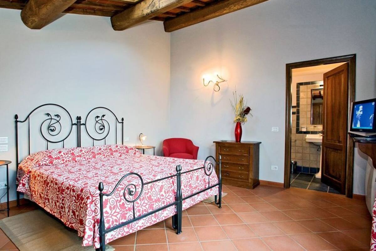 A bedroom with a bed, a red chair, a wooden dresser, a TV, and a wall-mounted light fixture.