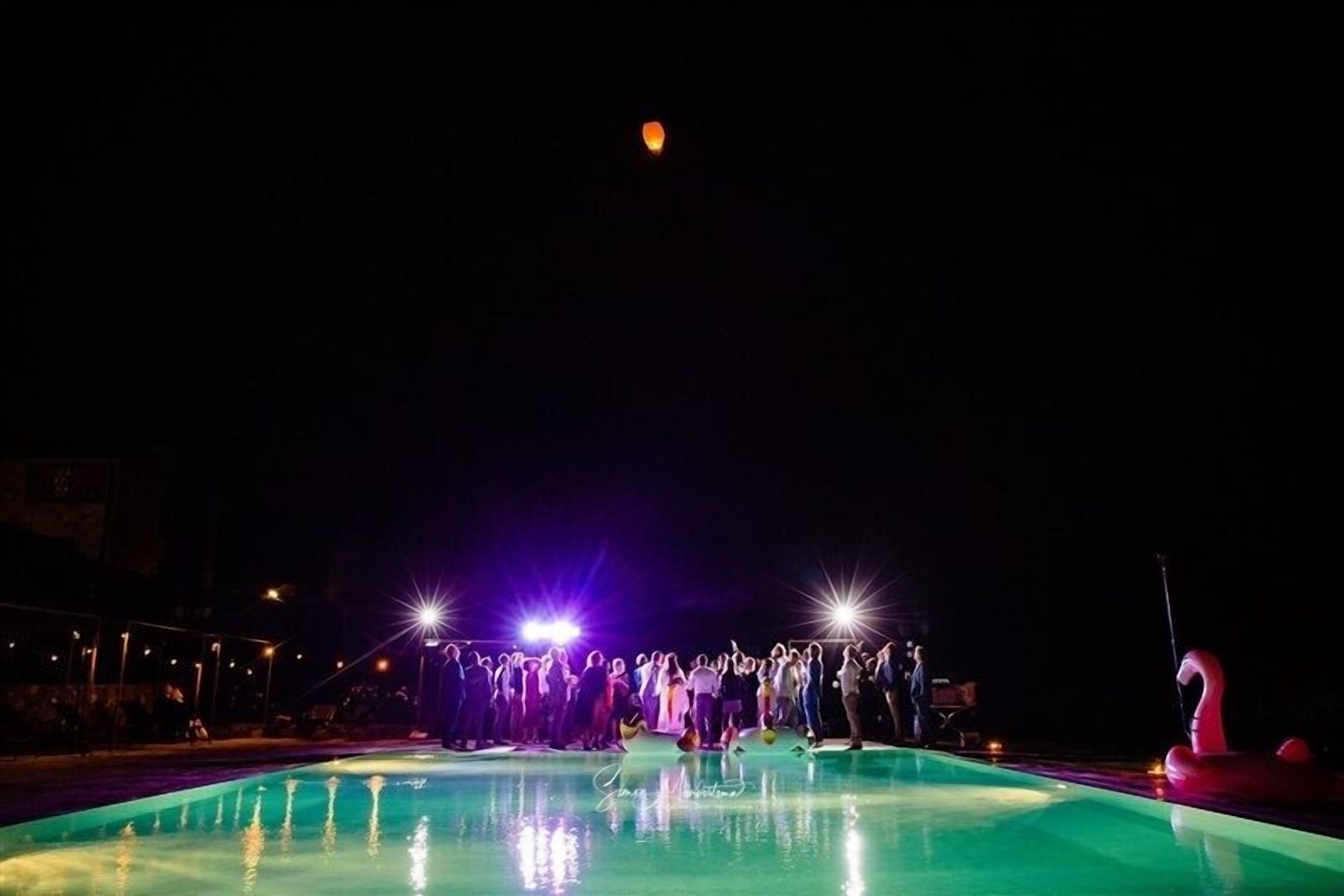 A nighttime outdoor pool area with people gathered around, illuminated by bright lights.