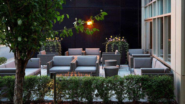 Een terras met buitenmeubels, waaronder banken en een vuurplaats, omgeven door groen en een gevel.