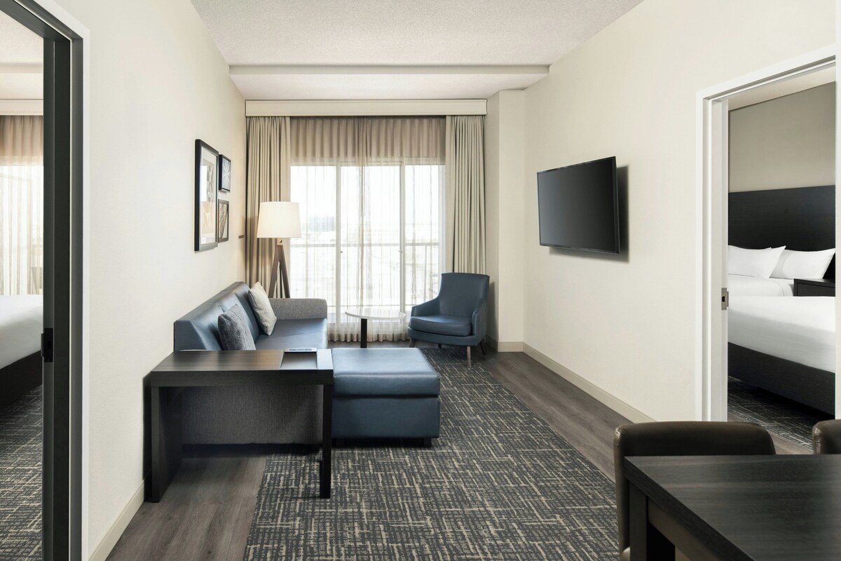Habitación de hotel con cama, sofá, sillón, televisión y ventana con cortinas.