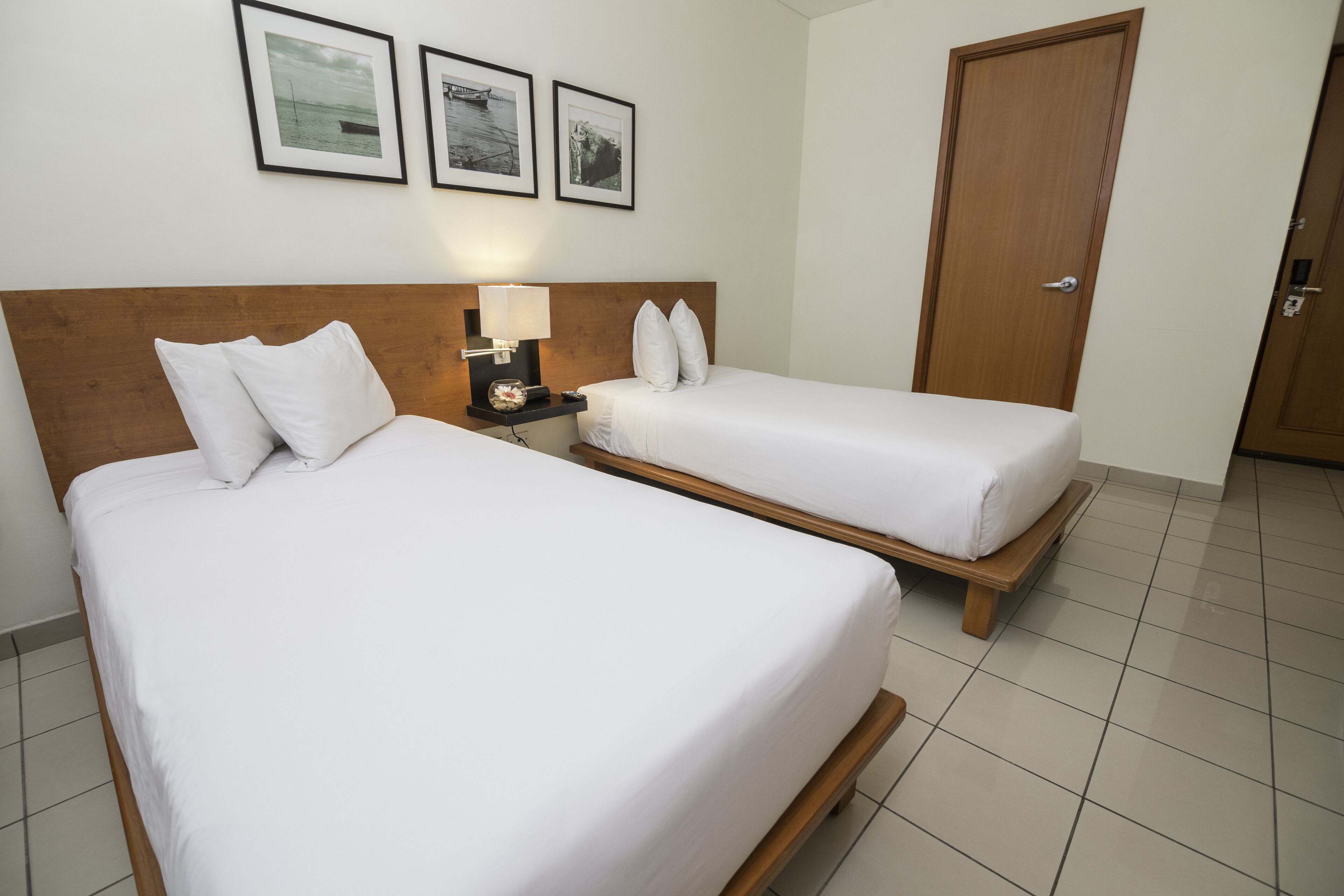 A hotel room with two single beds, a wooden headboard, a nightstand with a lamp, and a door.