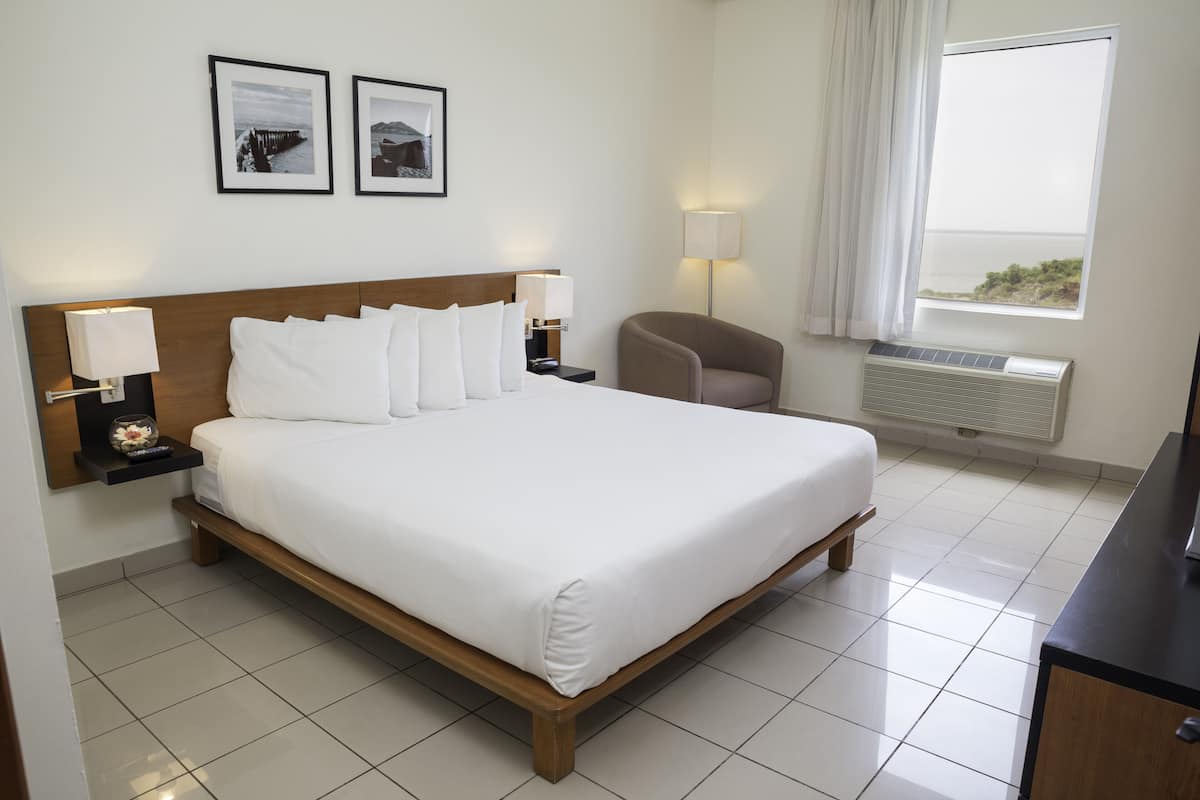 A hotel room with a large bed, two bedside lamps, a chair, a window with a view, and two framed pictures on the wall.