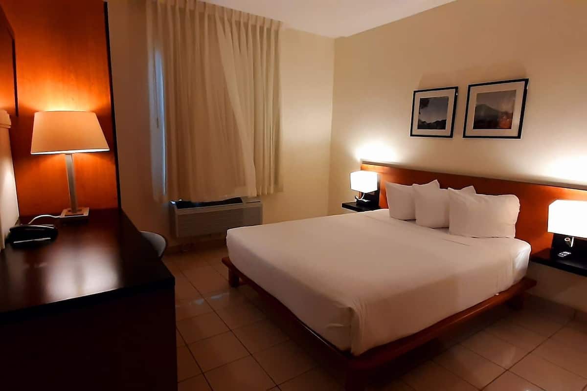 A hotel room with a bed, two bedside lamps, a desk with a lamp, a chair, a window with curtains, and two framed pictures on the wall.
