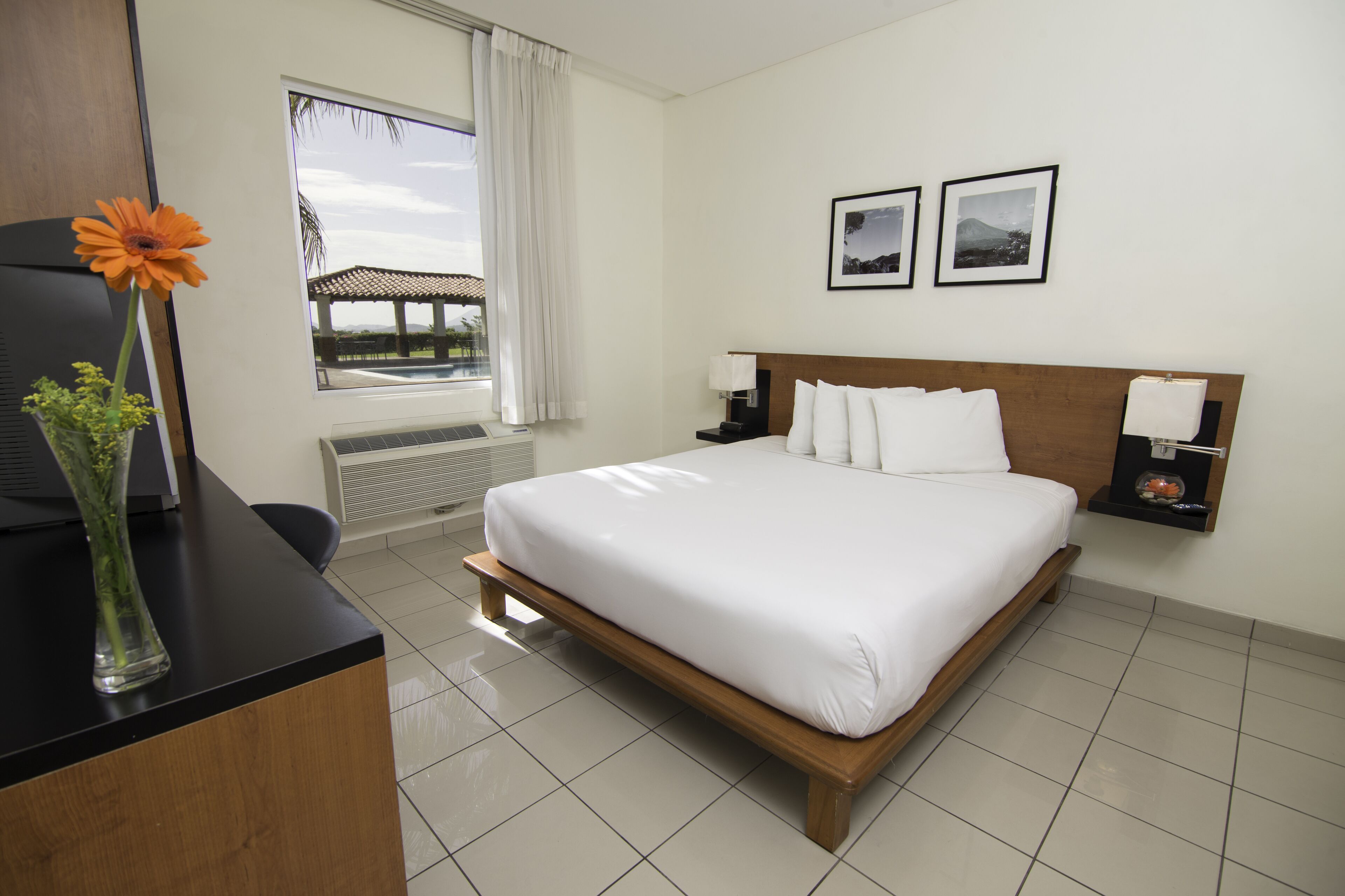 Habitación de hotel con cama, escritorio con un jarrón de flores, ventana con vistas y dos cuadros en la pared.