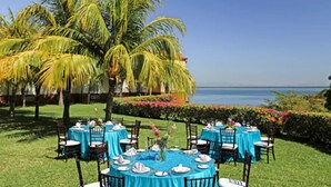 Un área de comedor al aire libre con mesas preparadas para una comida, rodeada de palmeras y vista al mar.